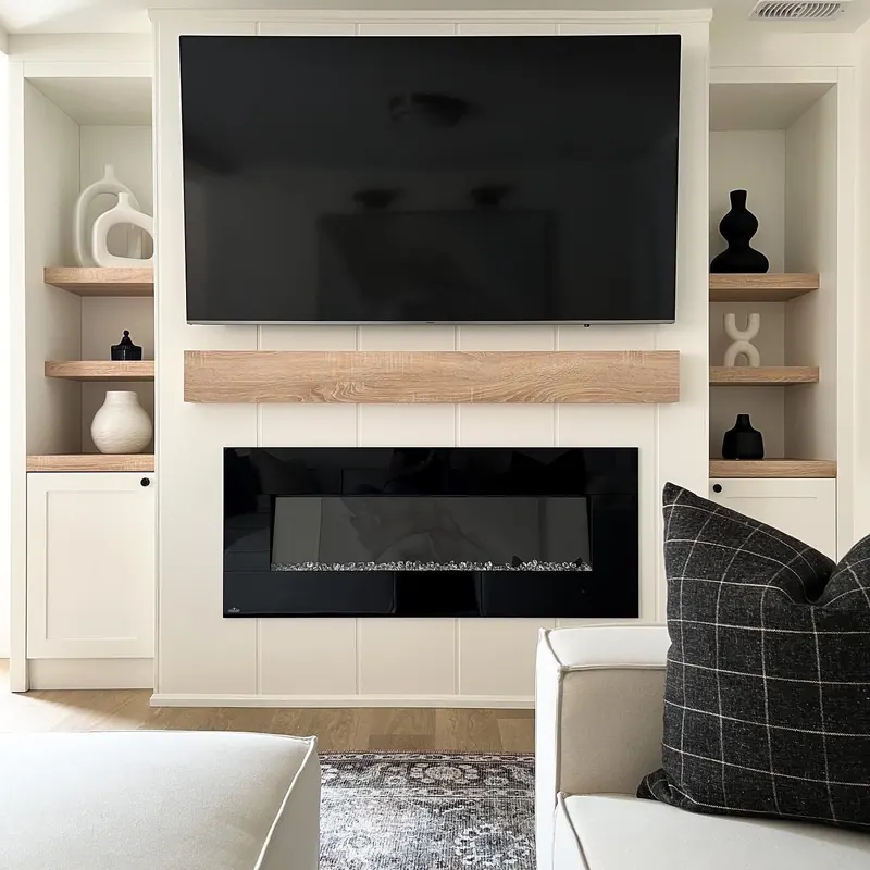 A media wall in white shiplap with an oak mantel, open shelves either side and a checked cushion in front