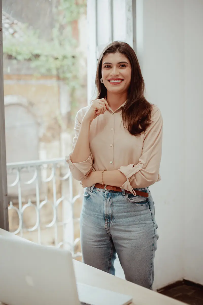 Érika Ferreira standing at a window, a laptop open on the desk in front of her